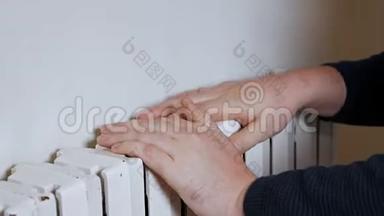 一个戴帽子的人把手放在靠近墙壁的暖气片上取暖。 公寓里冷，供暖系统差